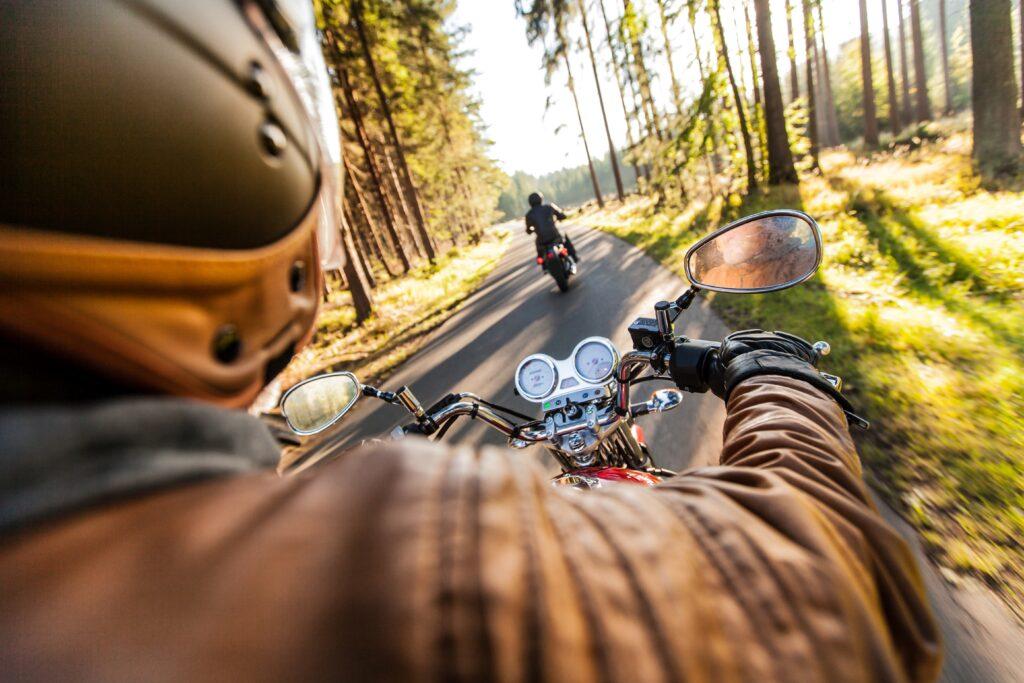 Blick über die Schulter eines Motorradfahrers, der auf einem 500 ccm Motorrad durch einen sonnendurchfluteten Wald fährt, mit einem anderen Motorrad im Hintergrund, umgeben von der Ruhe der Natur
