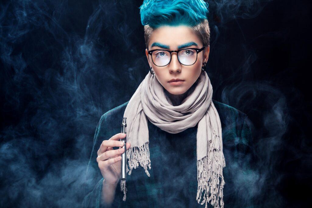 Person mit blauen Haaren und Brille, die eine Vape in der Hand hält, umgeben von Dampfwolken und stilvoll gekleidet mit Schal und kariertem Mantel.