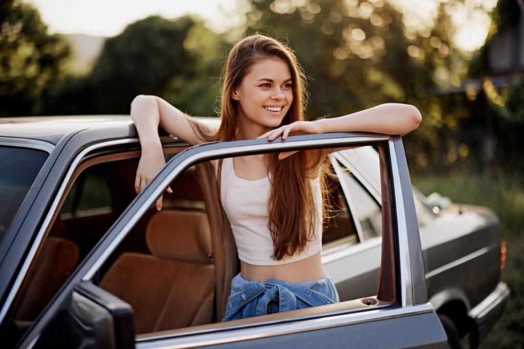 Junge Frau in weißem Top lehnt an der geöffneten Autotür eines Autos und lächelt, mit Sonnenuntergang und Natur im Hintergrund